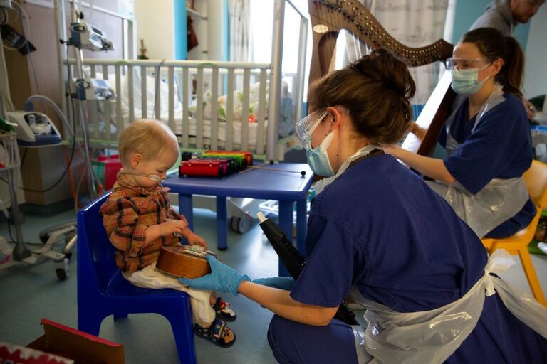 A harpist and other musician in a hospital room with a toddler playing on a ukulele