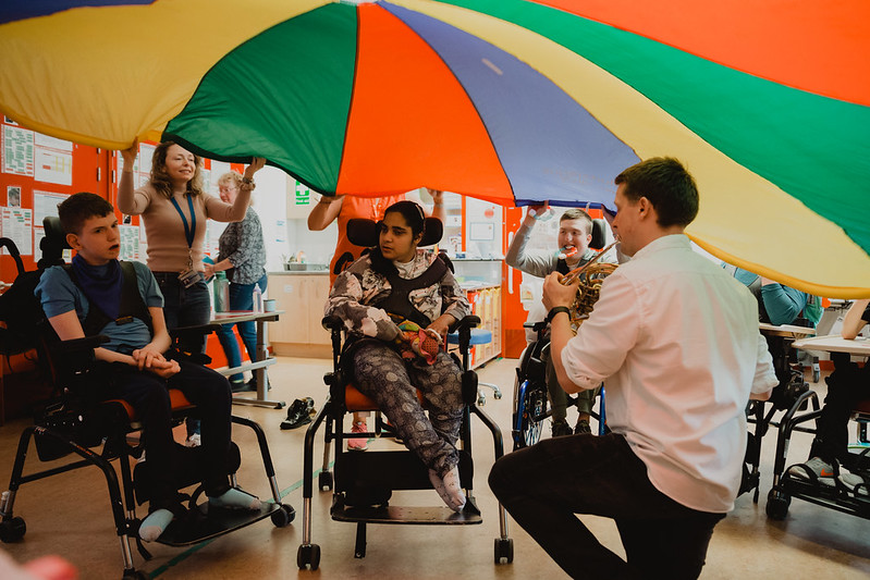 Three children in wheelchairs listening to musician playing french horn under a colorful parachute.