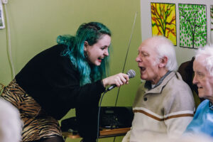 A young woman with teal hair holds a microphone for an older man who is singing, both smiling and enjoying a musical activity together.