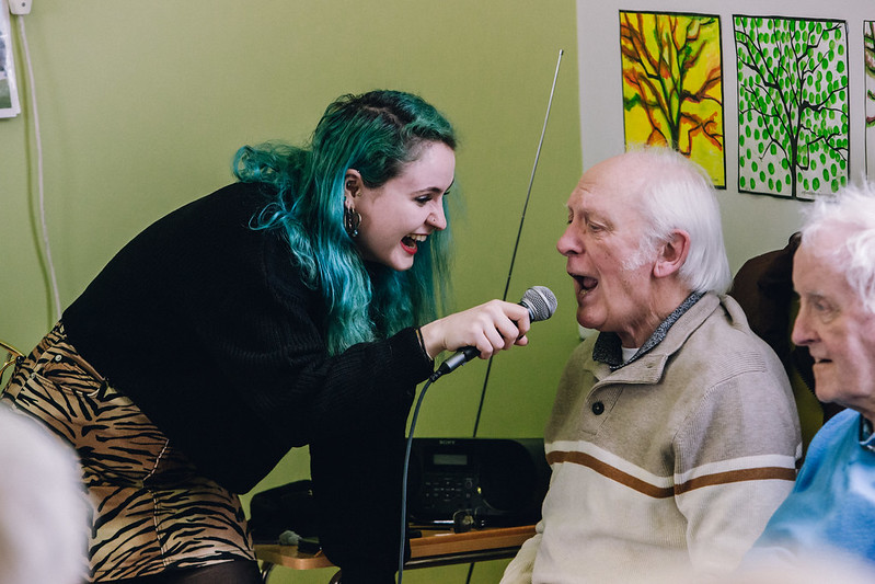 A young woman with teal hair holds a microphone for an older man who is singing, both smiling and enjoying a musical activity together.