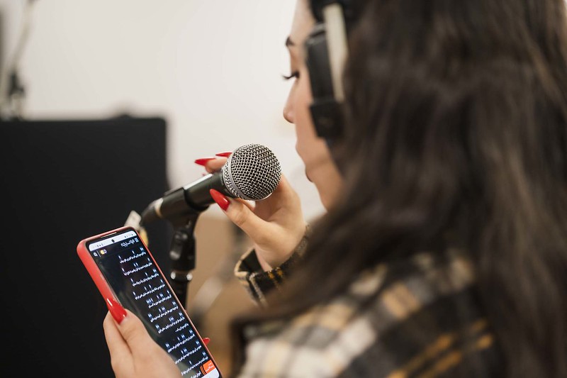 Young woman with headphones, singing into a microphone, looking at lyrics in Arabic on her phone.