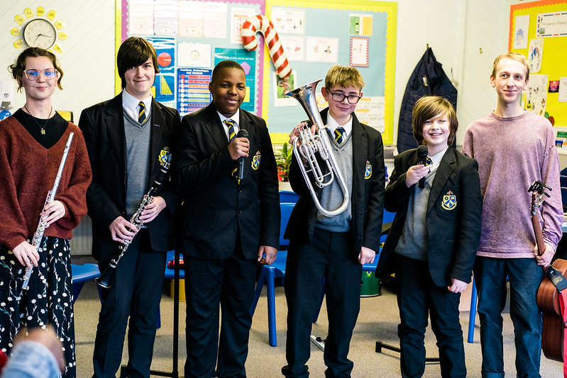 A group of school students in uniforms stand with their music teacher, holding instruments including a flute, clarinet, euphonium, guitar, and microphones.