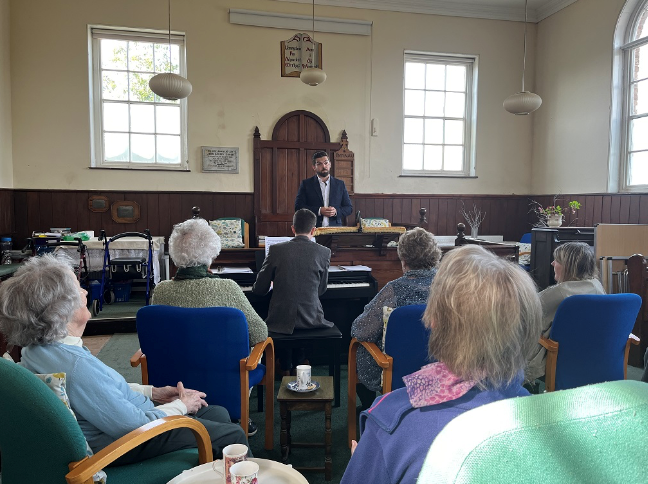 Man singing with piano accompaniment in front of an audience seated in a small hall.