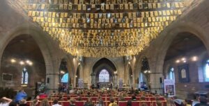 Church interior with congregation seated, golden cards hanging from the ceiling, and a stained glass window at the altar.