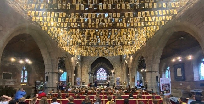Church interior with congregation seated, golden cards hanging from the ceiling, and a stained glass window at the altar.