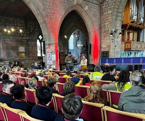Children and adults seated in a church, watching a live music performance at the front.
