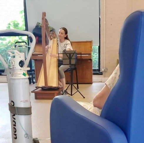 Woman playing a harp in a room, with a seated listener and medical oxygen tank visible in the foreground.