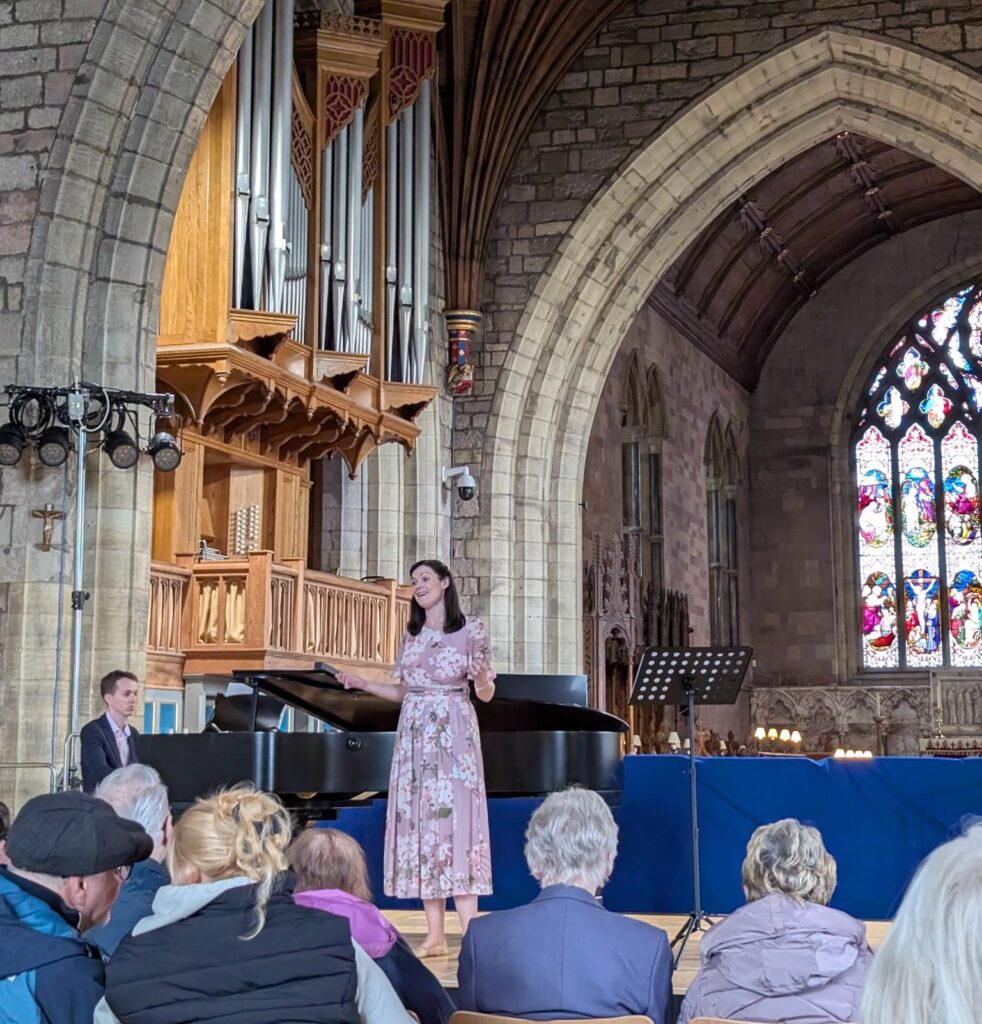Woman singing beside a grand piano in a church, with an organ and stained glass window in the background and an audience seated in front.