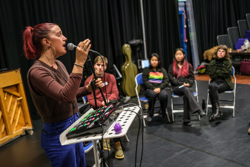 A musician singing into a microphone with electronic equipment, while other musicians watch.