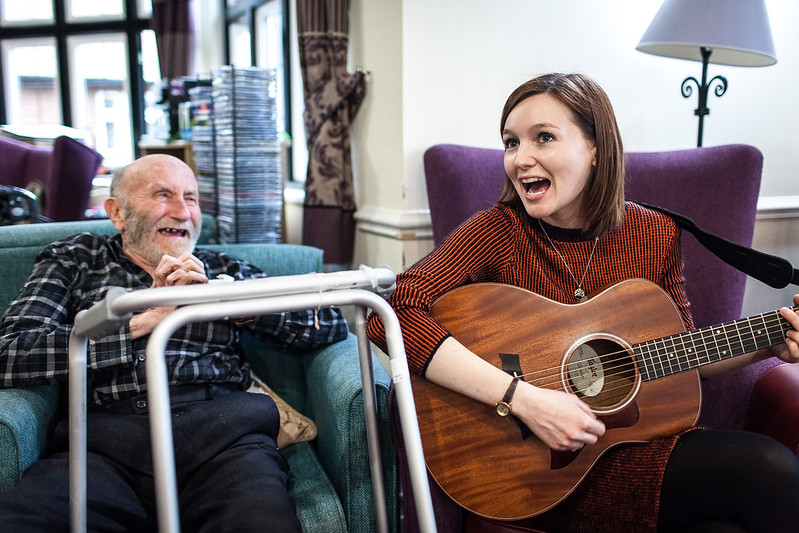 Elderly man smiling sitting next to musician playing guitar.