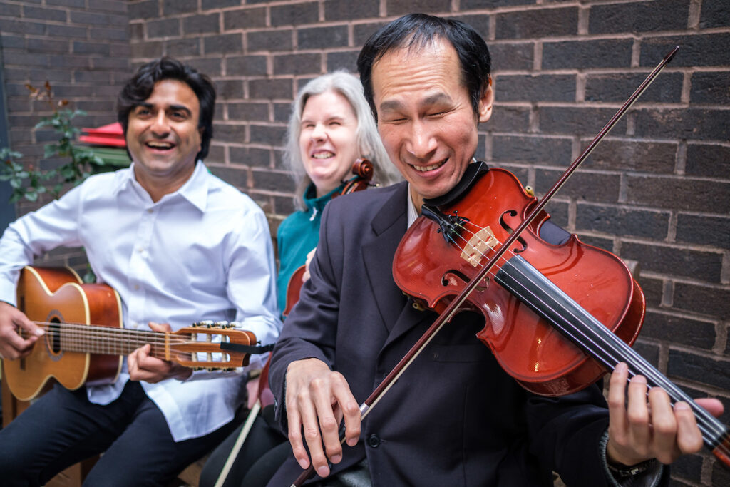 A man in a light blue shirt with a guitar is seated next to a woman with a violin and a man with violin.